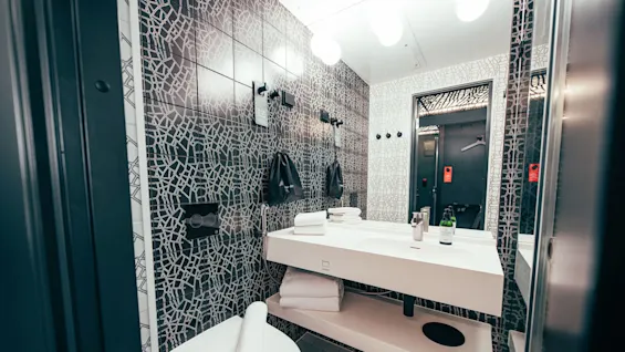 A modern bathroom features a white sink with a faucet, white towels, and an adjacent toilet. The walls have a black geometric pattern, and a mirror reflects more surroundings.