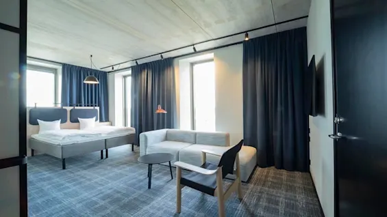 A modern hotel room with two beds, a light grey sofa, a chair, and dark blue curtains.