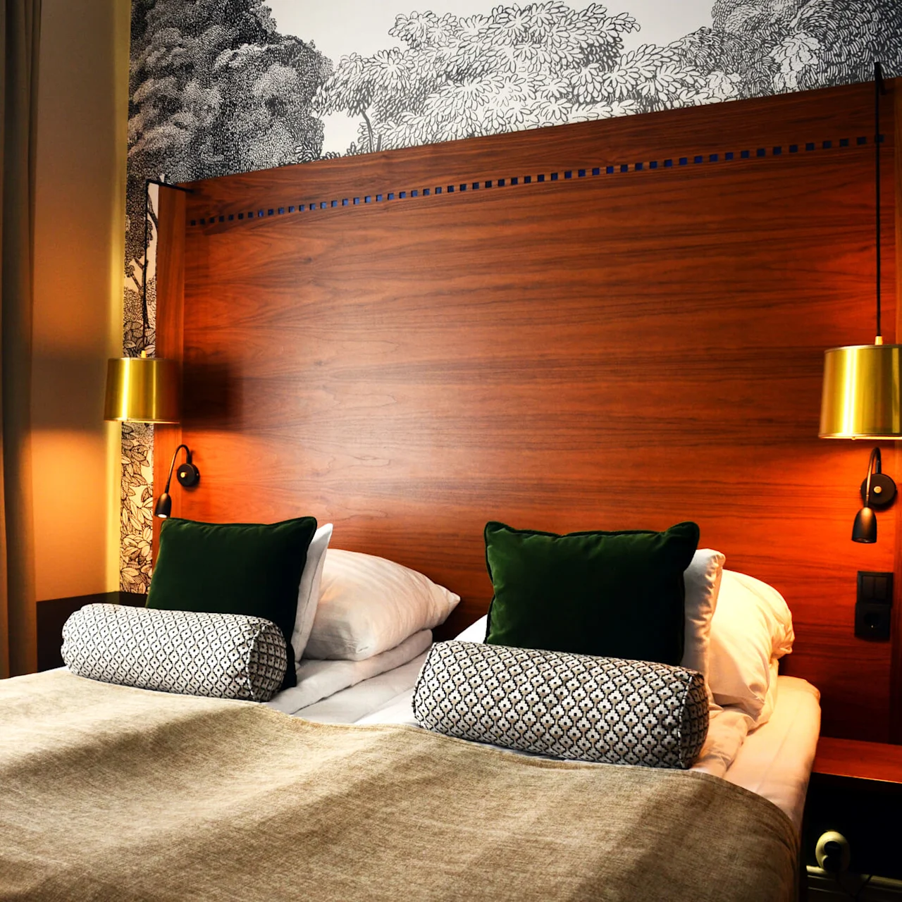 A cozy, neatly-made bed with pillows, flanked by brass wall lamps, against a wooden headboard and decorative wallpaper in a dimly lit room.