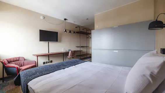 A neatly made double bed, covered in white linens and a gray blanket, stands in a modern hotel room with beige walls, featuring a wall-mounted TV, a pink armchair, and a desk with glass bottles.