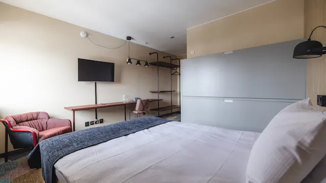 En snyggt bäddad dubbelsäng täckt med vita sängkläder och en grå filt står i ett modernt hotellrum med beige väggar, som har en väggmonterad TV, en rosa fåtölj och ett skrivbord med glasflaskor.