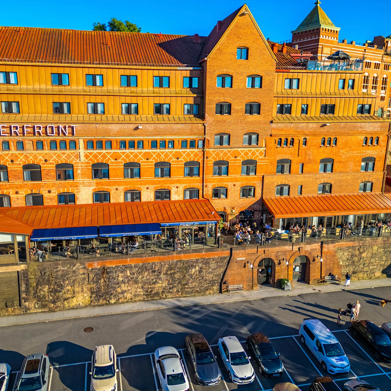 A large brick building labeled "WATERFRONT" features outdoor seating areas with people dining. It overlooks a parking lot filled with cars, under a clear blue sky.