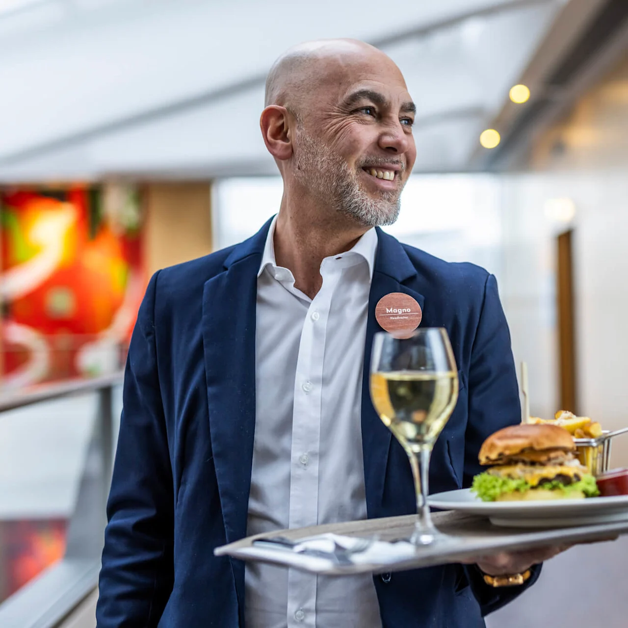 En man bär en bricka med en hamburgare, pommes frites och vin, leende i en ljust upplyst korridor. Han har på sig en namnbricka där det står "Magno".