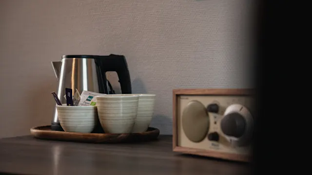 Electric kettle rests on a wooden tray with two ceramic mugs and tea sachets. Nearby, an old-fashioned radio sits on a dark wooden surface against a textured wall.