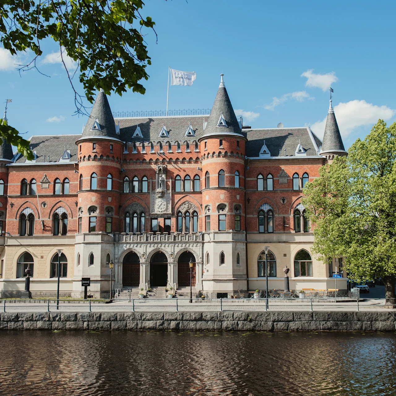 Storslagen byggnad med röda tegelstenar och välvda fönster, stående vid en flod under en klarblå himmel. Träd ramar in scenen och ger naturlig grönska.