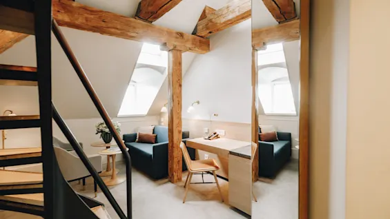 Ljust hotellrum på vinden med synliga träbjälkar, skrivbord, soffa och en trappa.