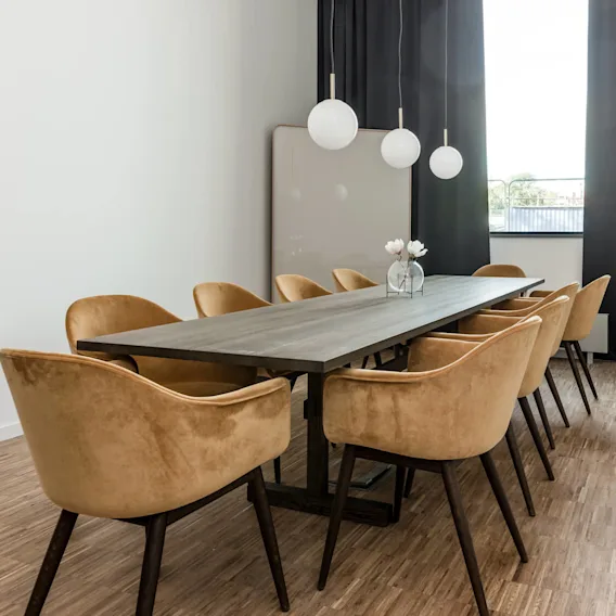 A long conference table with beige chairs surrounds it, set under three white pendant lights, in a room with wooden flooring and a large window with black curtains.