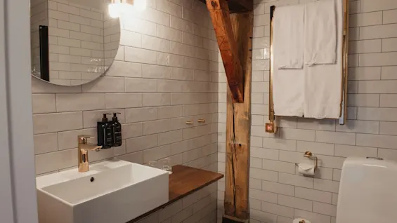 Et moderne badeværelse med hvide fliser, en vask med guldfarvede armaturer og en rustik træbjælke. Hvide håndklæder er pænt foldet.