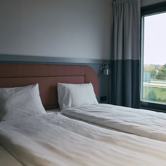 Ett mysigt hotellrum på Stopover Hotel Falkenberg med två sängar och ett stort fönster med utsikt över en väg och grönt landskap.