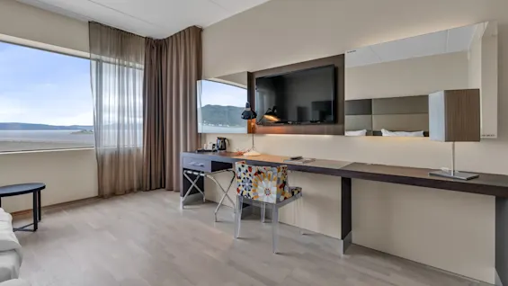 TV on a wall-mounted shelf above a desk with a lamp and chair. Large window with curtains reveals ocean view. Text on mirror: "LOOK GREAT!".