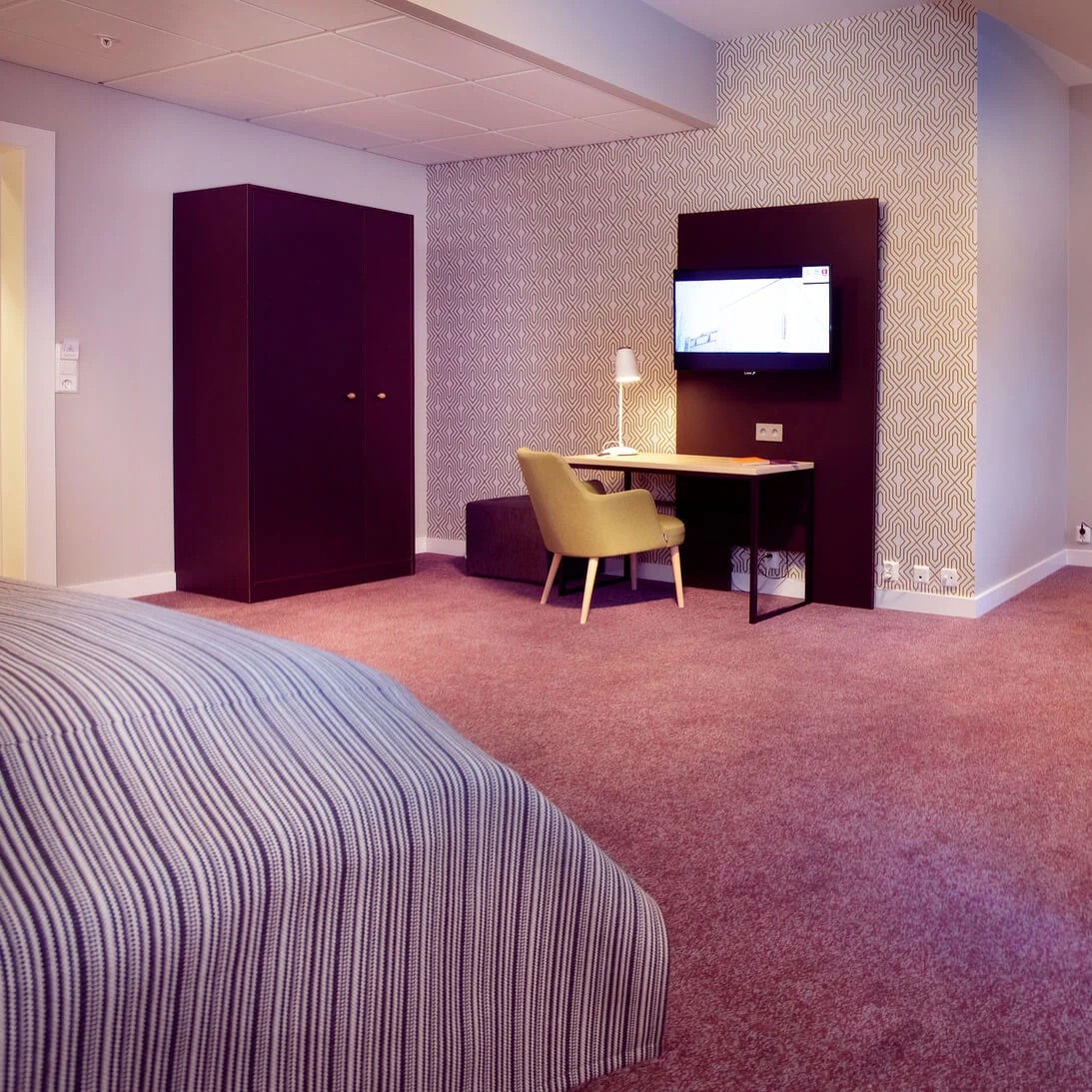 Et ryddig moderne hotellrom med mønstret vegg, en seng, stoler, et skrivebord, et klesskap og lamper, som viser komfortable møbler i et koselig, varm belyst rom.