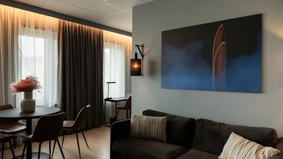 Et koselig hotellrom med en mørk sofa, et spisebord, et skrivebord og et stort abstrakt maleri på veggen.