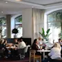 People sit and converse around tables in a warmly lit lounge area. The space features large windows, indoor plants, and modern furniture, creating a relaxed, inviting atmosphere. 