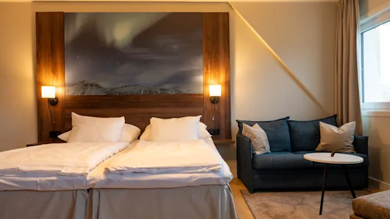 Et koselig hotellrom med to senger og en sofa, med et sengegavlmaleri av nordlyset over snødekte fjell.