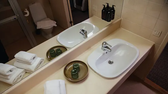 A white sink reflects on a mirror above a beige countertop. Rolled towels and small glass containers sit nearby. A toilet is visible in the background.