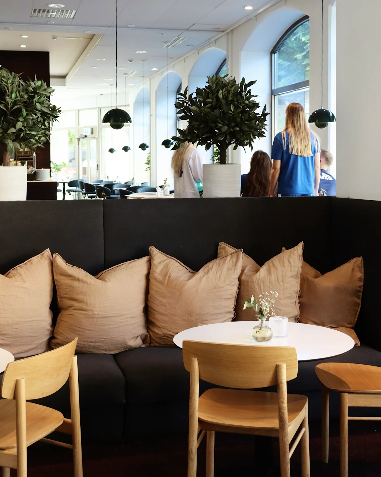 Comfortable seating area with beige cushions and a small round table, surrounded by wooden chairs. Two potted plants are placed on the partition. People stand near large windows in the background.