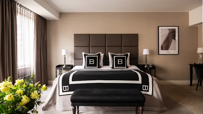 Seng med sort og hvidt interiør, med puder med bogstavet "B", står i et moderne rum med neutrale toner. Et vindue med gardiner, indrammet kunst og en vase med blomster øger elegancen.