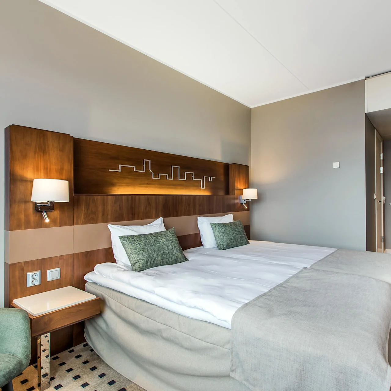 A neatly made hotel bed with green pillows rests against a wooden headboard with skyline design, beside a green armchair and wooden furniture in a modern, softly lit room.