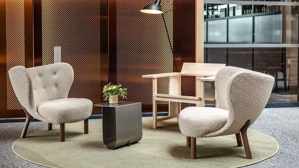 Comfortable cream chairs and a wooden armchair rest on a circular rug with a small metal table in between; a potted plant sits on the table, surrounded by a perforated wall and a floor lamp.