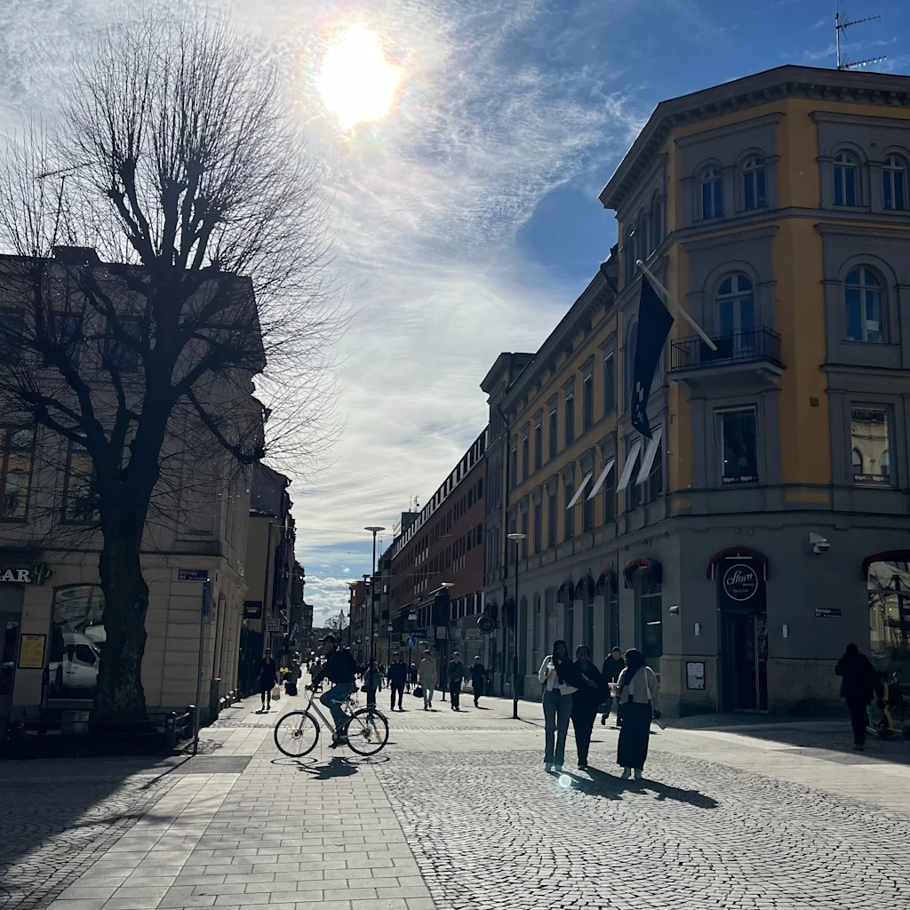 A sunny street scene with people walking and cycling on a paved and cobblestone street, flanked by buildings, one with a 'TACO BAR' sign.