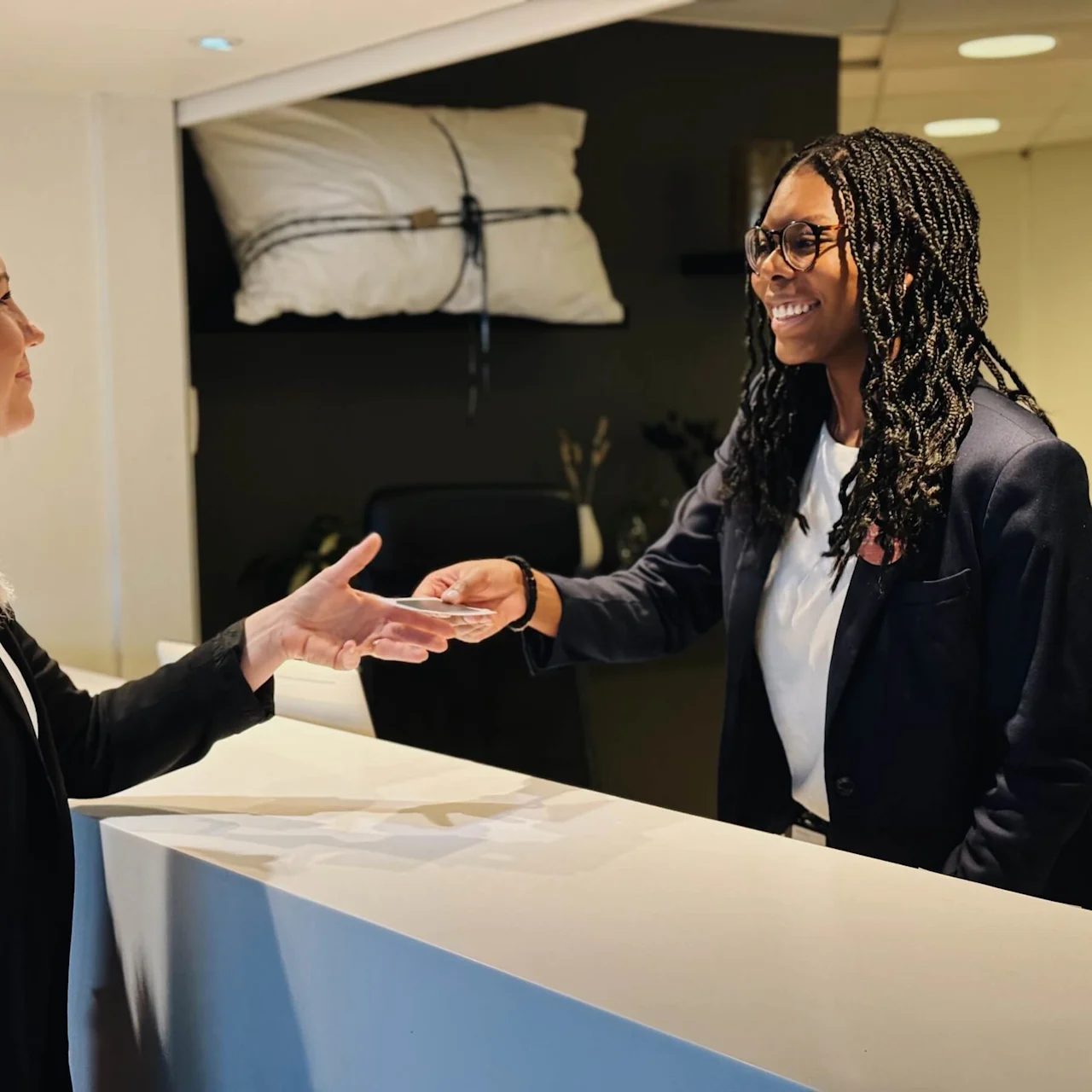 A woman hands an ID card to a smiling receptionist at a hotel front desk. A decorative pillow hangs in the background, and the area is well-lit.