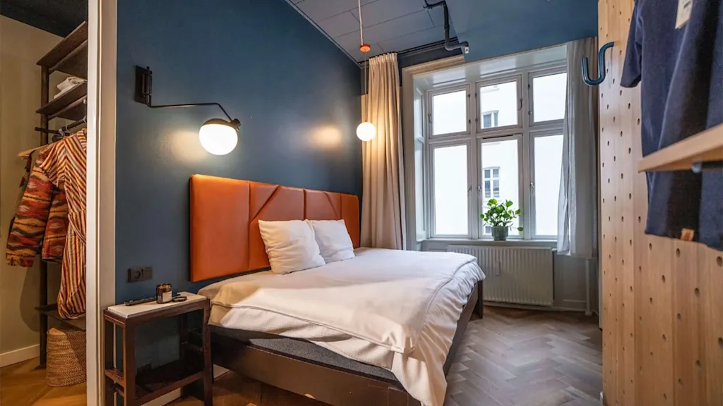A modern hotel room with a double bed, blue walls, wooden floors, and a large window letting in natural light.