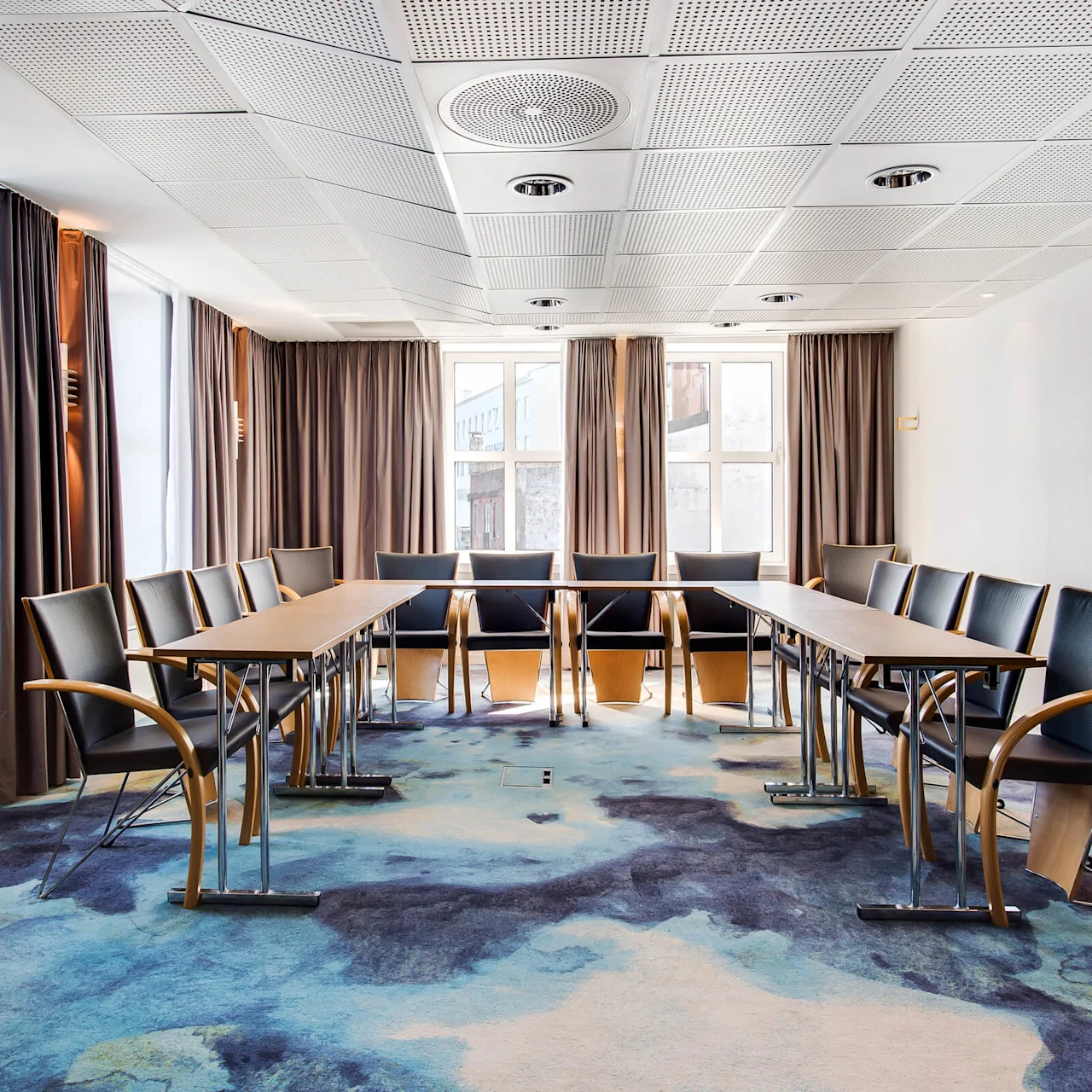 Et konferencelokale har en U-formet bordopsætning med sorte stole, omgivet af store vinduer og brune gardiner, under et loft med indbygget belysning. Et mønstret blåt tæppe dækker gulvet.