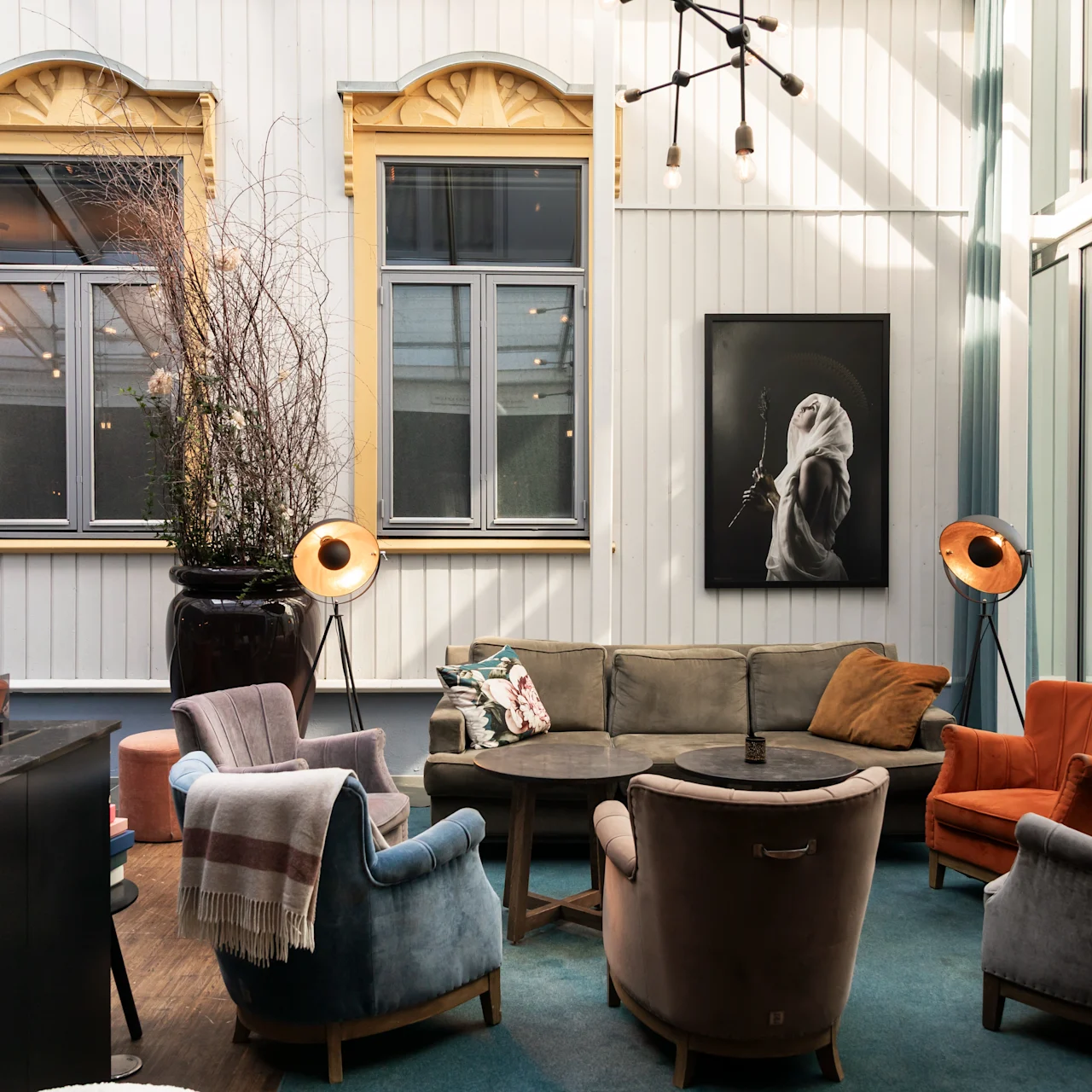 Komfortable stole og sofaer omringer et rundt bord i et hyggeligt, stilfuldt rum med høje vinduer og dekorativ vægkunst; blød belysning og neutrale toner skaber en varm, indbydende atmosfære.