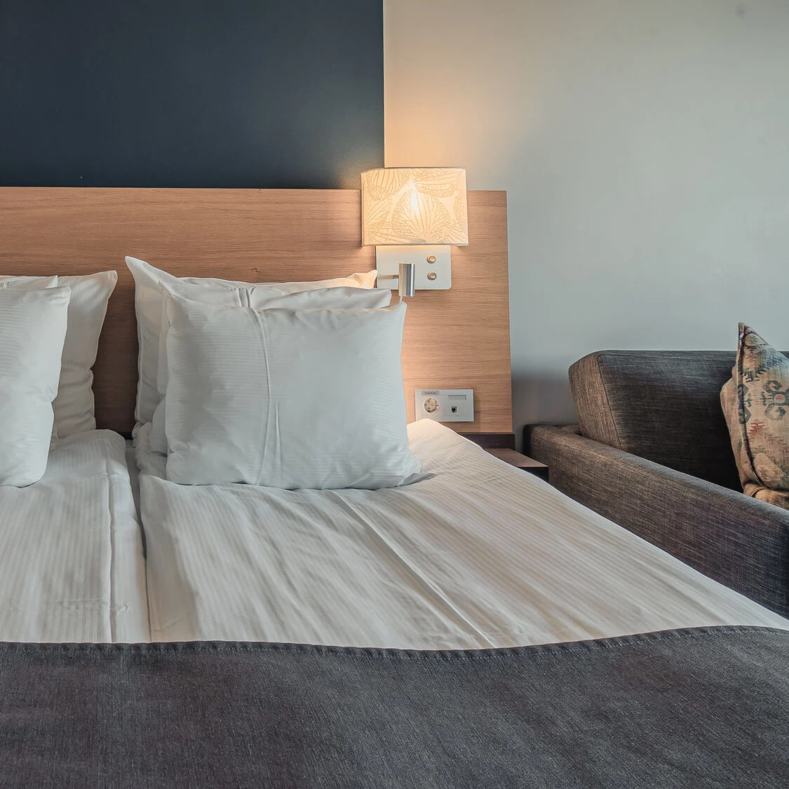 A double bed with white sheets and pillows is neatly arranged in a modern room. Two wall lamps illuminate the wooden headboard. A dark sofa is partially visible on the right.