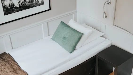 En pænt redt enkeltseng med en grøn pude, op mod en panelvæg, ved siden af et sengebord og en lampe.