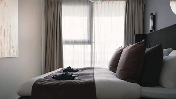 A neatly made bed with brown cushions and folded slippers rests in a softly lit room. Sheer curtains cover the window, and a nature-themed painting hangs on the wall.