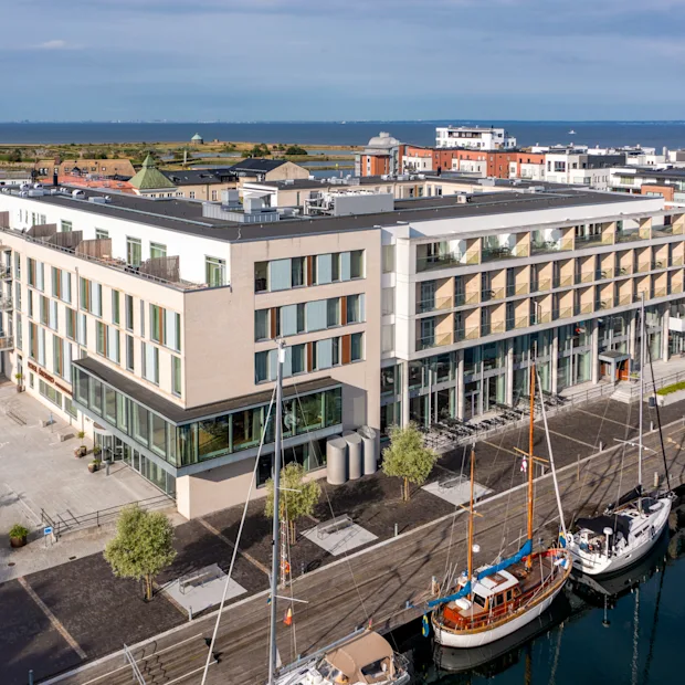 Moderni hotellirakennus lasijulkisivulla, josta on näkymä satamaan, jossa on useita veneitä, kaupunki ja meri taustalla.