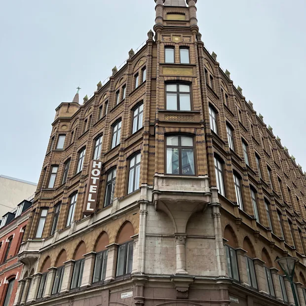 En murbygning med flere etasjer viser ordet "HOTELL" vertikalt på fasaden, og har butikker merket "Mulberry" og "ETON" på bakkeplan.