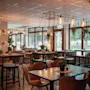 A cozy restaurant or cafe interior with a mix of high and low tables, warm lighting, and large windows overlooking greenery.