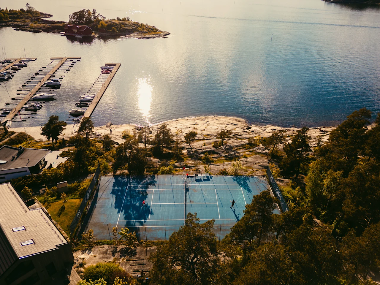 Et luftfoto af en tennisbane ved vandet, med folk der spiller, omgivet af træer og en marina med både.