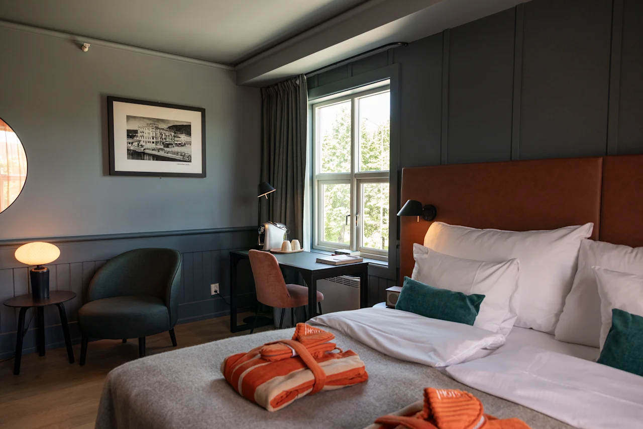 En prydligt arrangerad hotellrum har en säng med vita lakan och orange morgonrockar, bredvid ett fönster. Ett skrivbord med lampa och stol ger arbetsutrymme intill sängen.