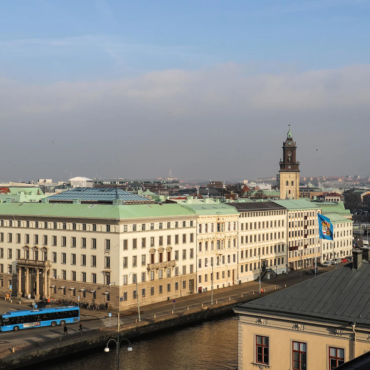 En stor, historisk byggnad med ett klocktorn blickar ut över en stadskanal. En blå buss passerar framför, och en flagga vajar i närheten under en delvis molnig himmel.