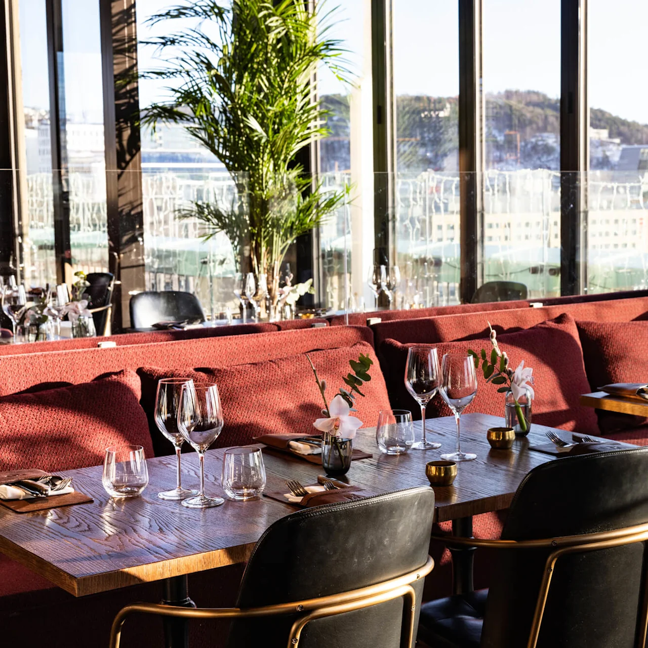 Tables set with wine glasses and cutlery rest against red cushioned seating in a sunlit restaurant with large windows and a view of city buildings and hills.