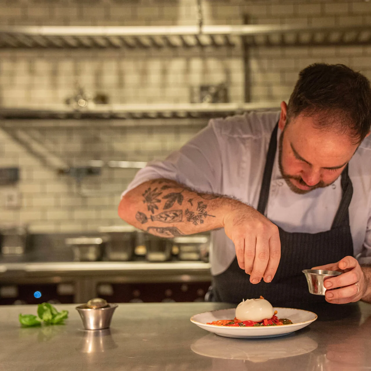 A chef decorates a dish with herbs, leaning over a stainless steel counter in a professional kitchen lined with white subway tiles and shelves.