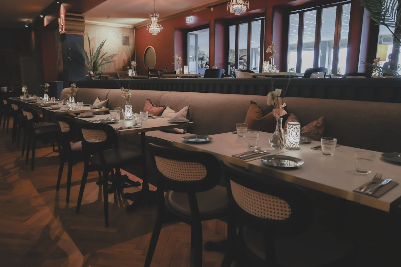 A long table is elegantly set with cutlery, glasses, and decorative flowers; chairs are neatly aligned. The restaurant interior features warm lighting and comfortable seating.