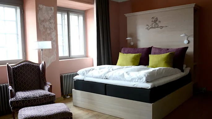 A bed with purple and green pillows is set against a wooden headboard with lamps; nearby is an upholstered armchair and ottoman. The room has large windows and wooden flooring.