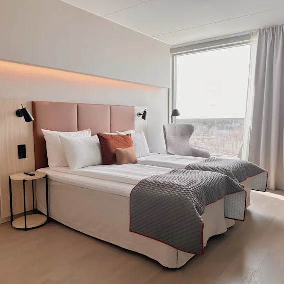 En säng med vita kuddar och grått överkast står bredvid ett litet runt bord och en modern grå stol. Dagsljus strömmar in genom stora fönster och lyser upp det minimalistiska rummet.