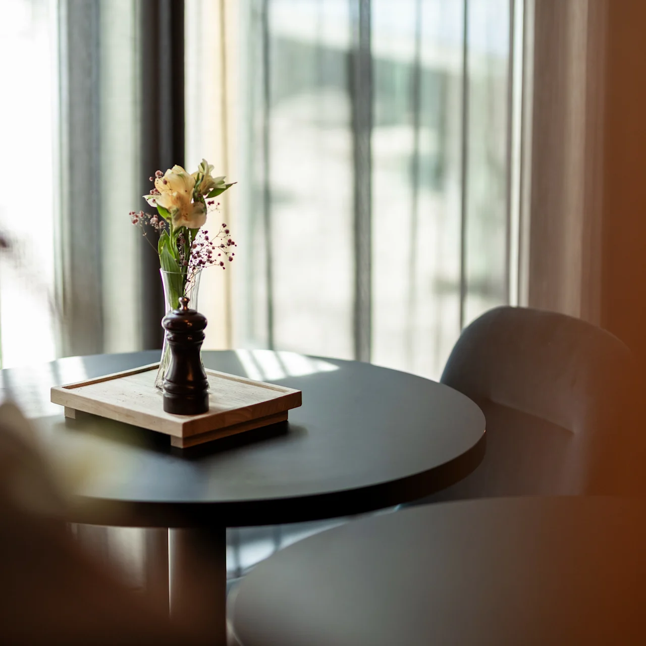 Et rundt bord har en lille vase med blomster og en peberkværn på en træbakke i et blødt oplyst rum med gennemsigtige gardiner.