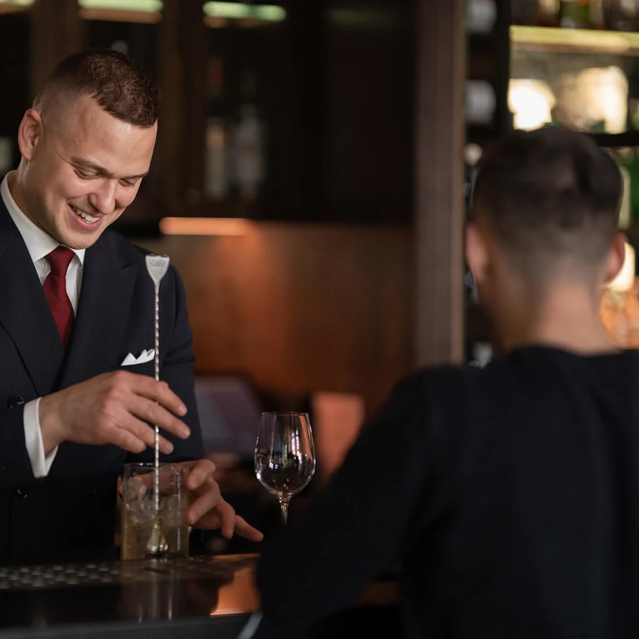 A bartender in a suit stirs a drink with a long spoon, smiling at a customer. They are in a dimly lit bar with bottles and glasses in the background.