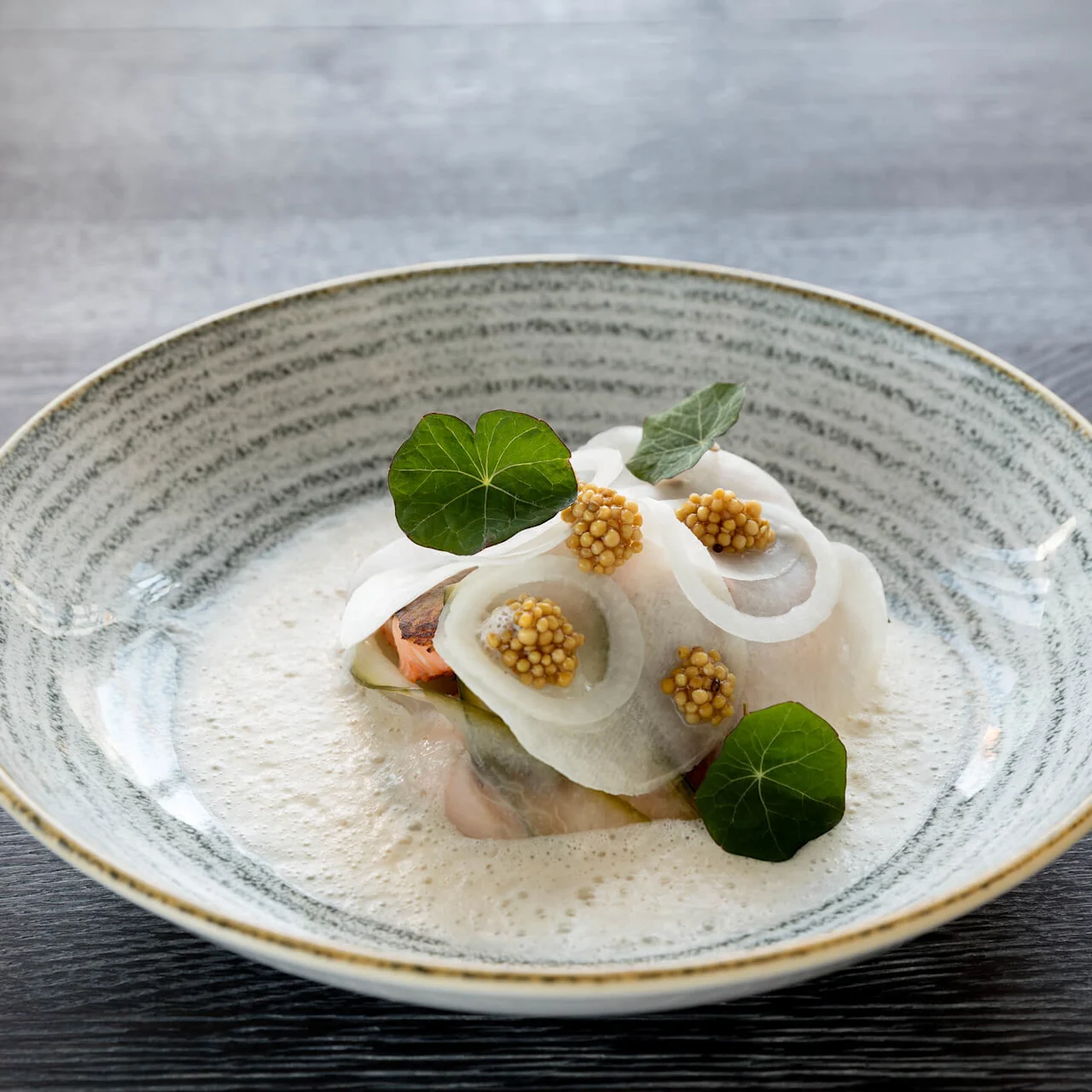 En rett med tynne skiver av reddik på toppen av en kremet saus, pyntet med sennepsfrø og grønne blader, elegant plassert i en flekkete skål på en mørk treflate.