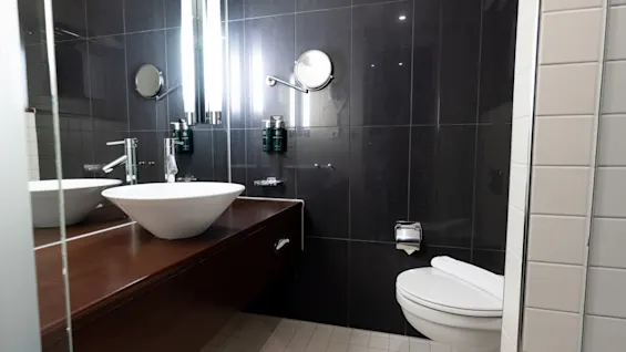 A modern bathroom features a round basin on a wooden countertop beside a wall-mounted toilet. On the dark tiled wall, there are mirrors and dispensers under bright lights.