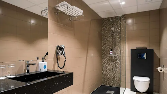 Et moderne badeværelse har en håndvask med armatur, sæbedispenser og hårtørrer, ved siden af en brusekabine og toilet, med brune flisevægge.