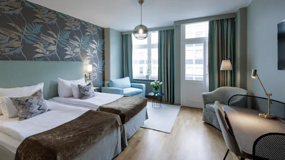 Välutrustat hotellrum med två enkelsängar, en fondvägg med lövdesign, möbler och fönster som släpper in naturligt ljus. Rummet känns bekvämt och stilfullt.