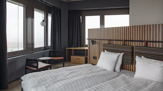 A neatly made bed with striped sheets sits under a wooden slatted headboard in a modern hotel room. Large windows provide natural light, complemented by a small seating area.