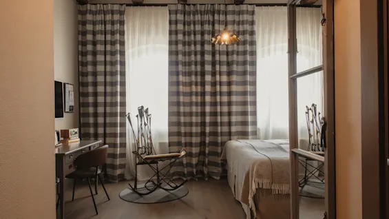 A cozy hotel room with a bed, a desk, and a unique rocking chair. Striped curtains cover large windows, and wooden beams adorn the ceiling.
